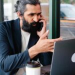 Bearded businessman multitasking with phone call and laptop in modern cafe setting.
